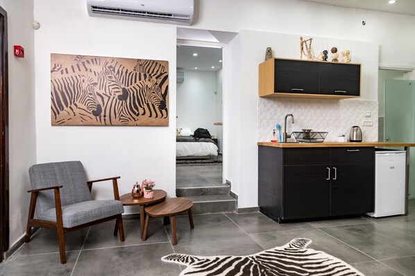 A modern living room with a grey armchair, a small round table, and a zebra-patterned rug. A kitchen with black cabinets and a white countertop is visible in the background.