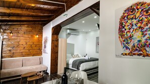 A cozy room with a wooden ceiling, a sofa, a dining table with glasses and a bottle, and a bed with a colorful artwork on the wall. - Kinneret Lodging (Tiberias)