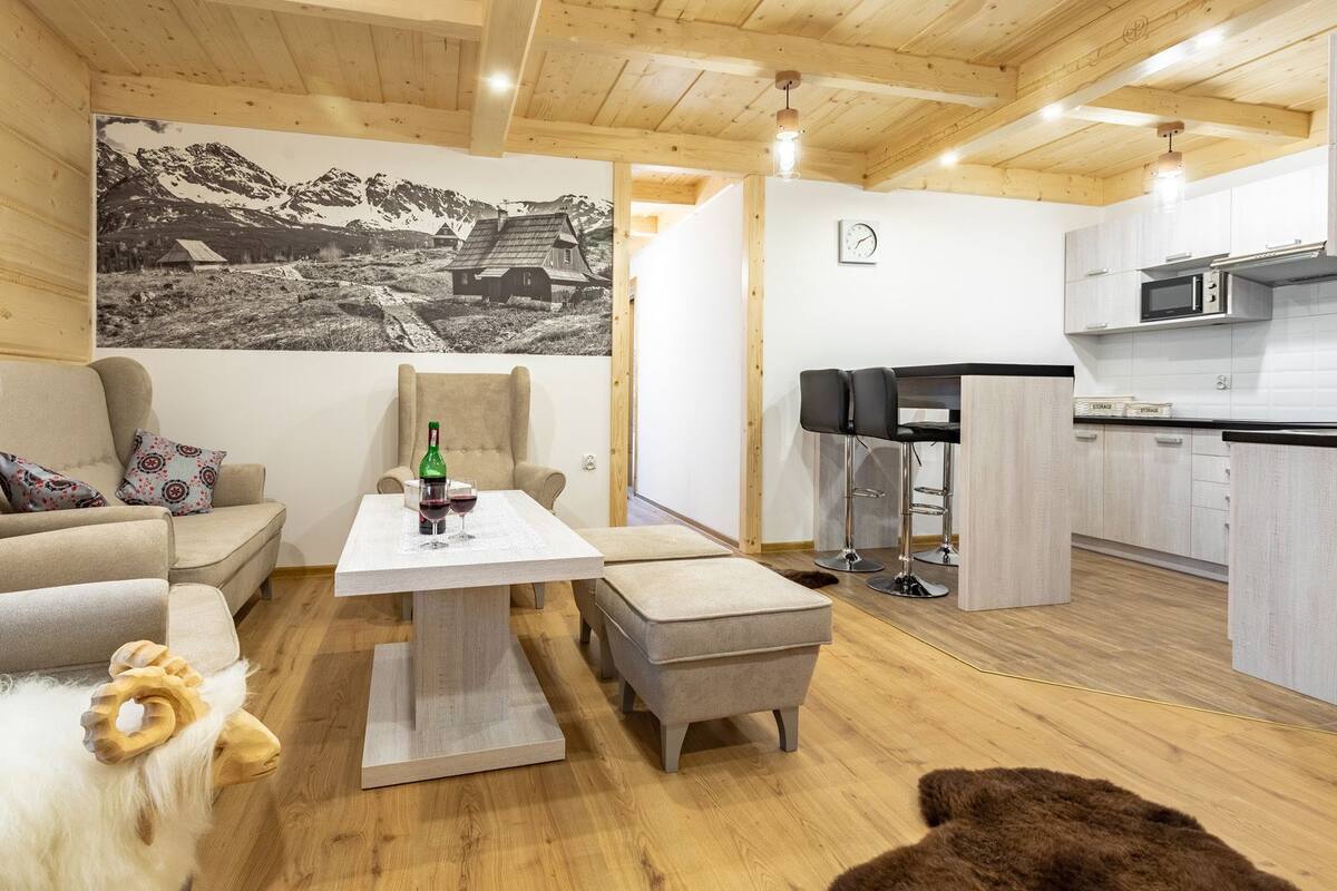 A cozy living area with a sofa, a table with wine glasses, a chair, and a kitchen with a microwave.