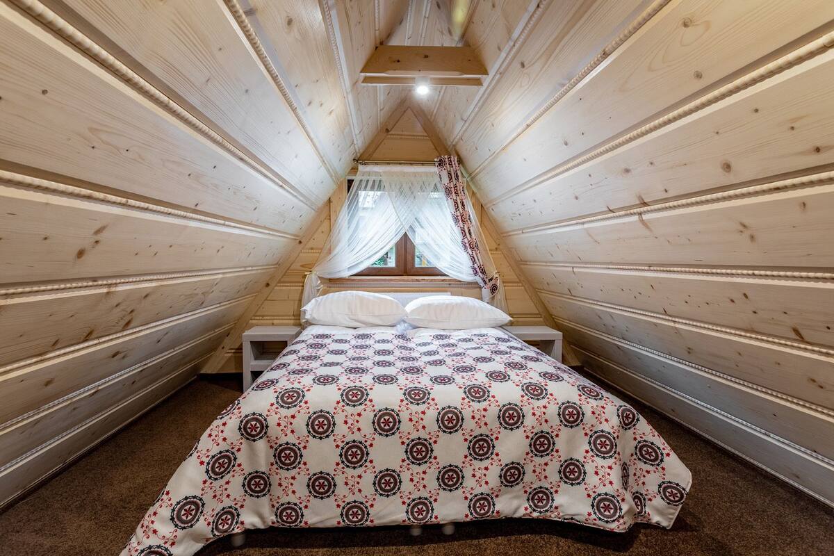 A cozy attic bedroom with a sloped ceiling, a bed with a patterned bedspread, and a window with sheer curtains.