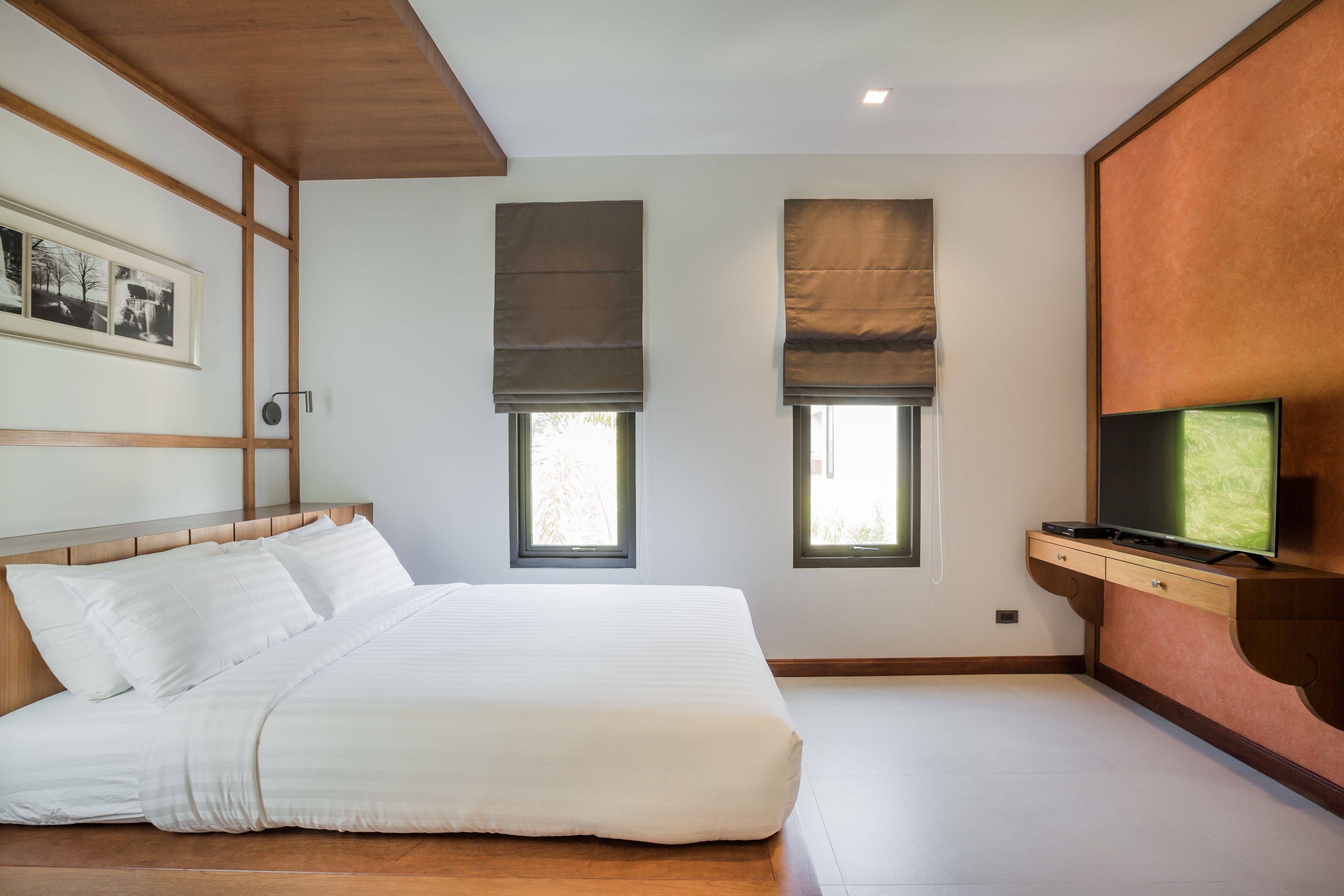 A bedroom with a bed, two windows with blinds, a TV on a wooden cabinet, and a framed picture on the wall.