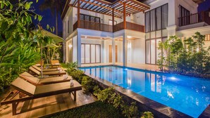 Una casa de dos pisos con piscina, terraza de madera con sillas de descanso y un patio cubierto.