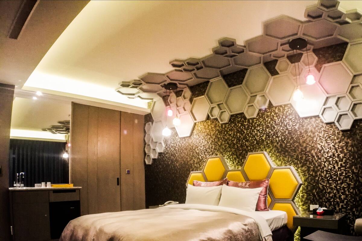 A modern hotel room with a unique hexagonal ceiling design, a bed with a yellow headboard, and a small bedside table.