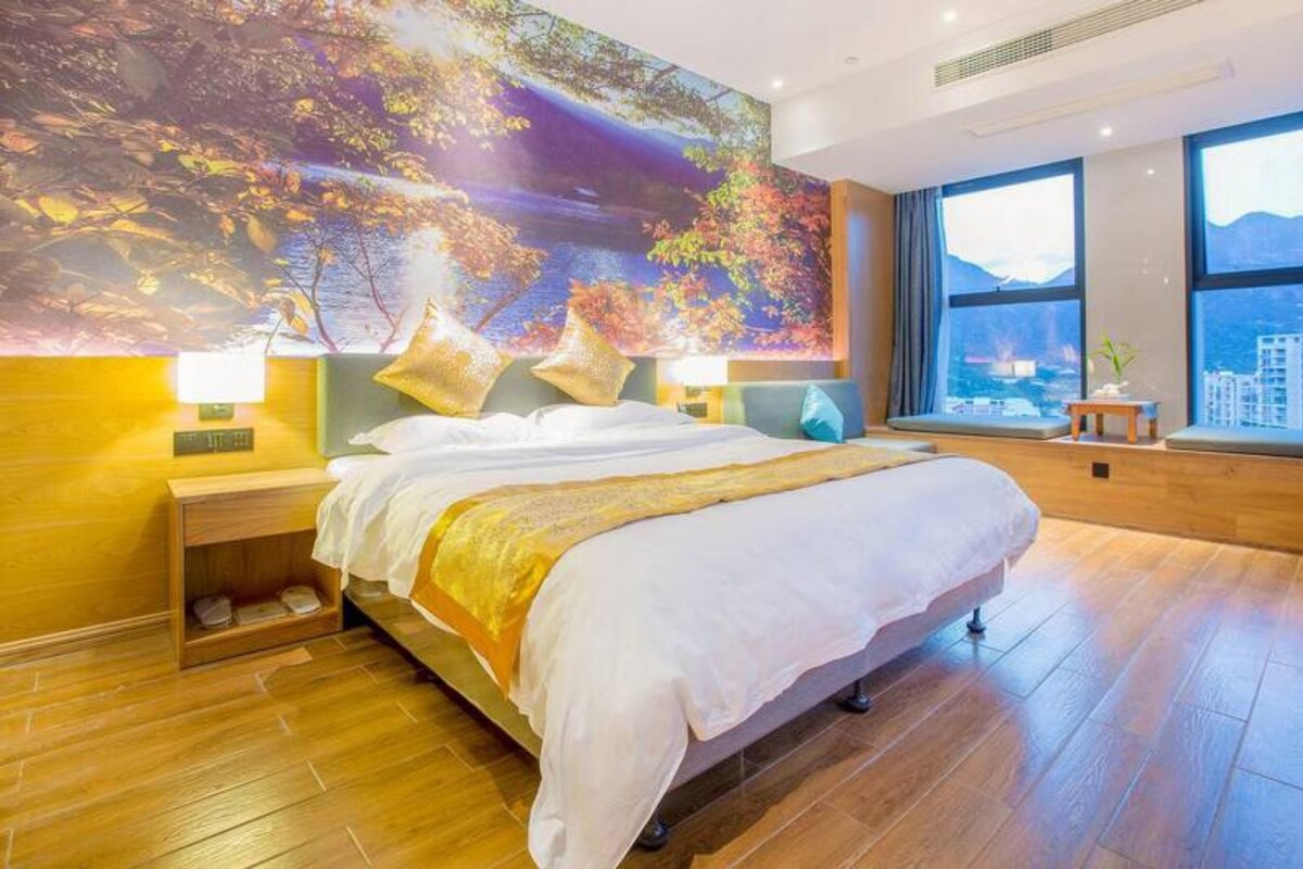 Ein geräumiges Schlafzimmer mit einem großen Bett, Blick auf die Stadt durch große Fenster und einem Landschaftsgemälde an der Wand.