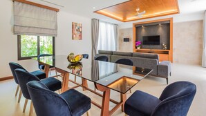 A modern living room with a glass dining table, dark blue chairs, a grey sofa, and a flat-screen TV mounted on the wall. - Villa Palavee B3 (Krabi)