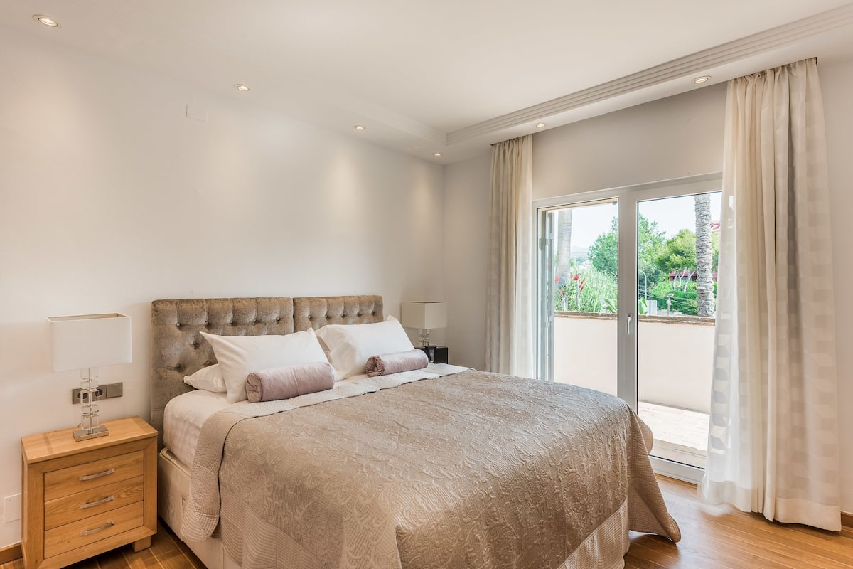 Une chambre avec un grand lit, une table de chevet, une lampe et une vue sur la verdure par la fenêtre.