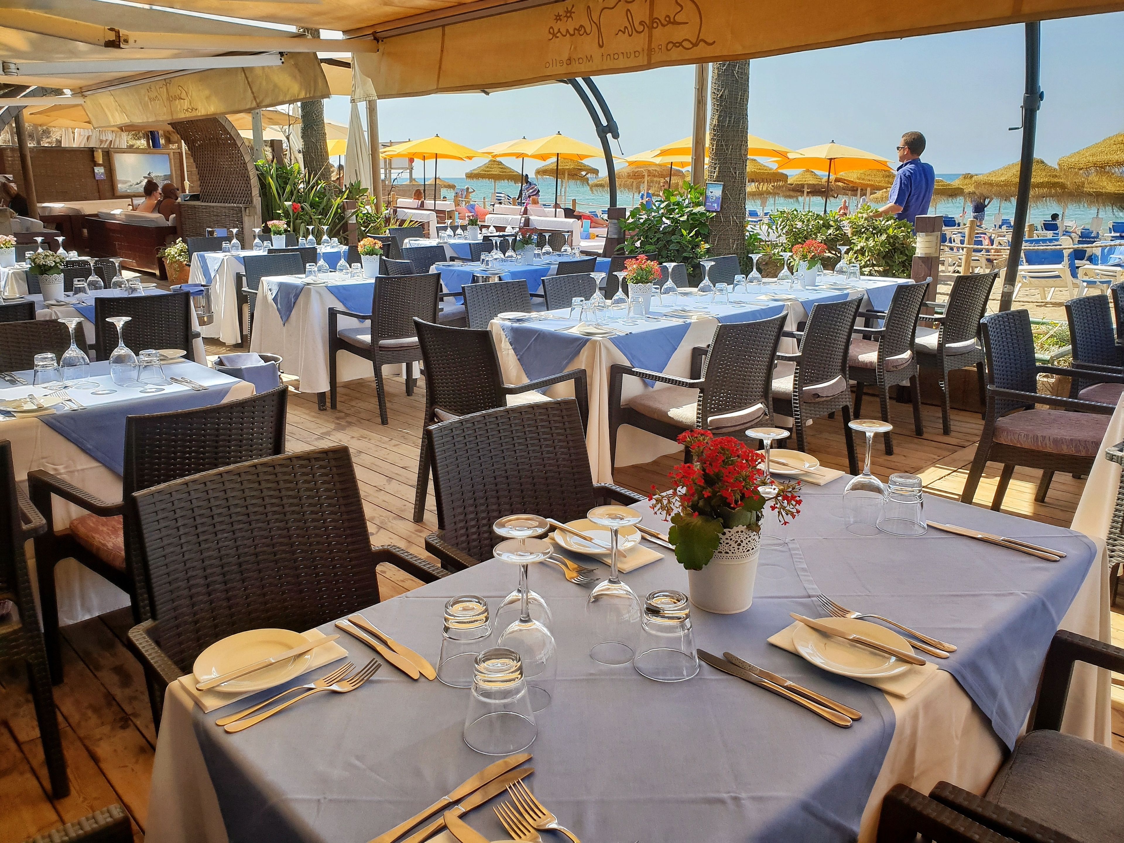 An outdoor dining area with tables set for two, featuring wicker chairs and blue tablecloths.