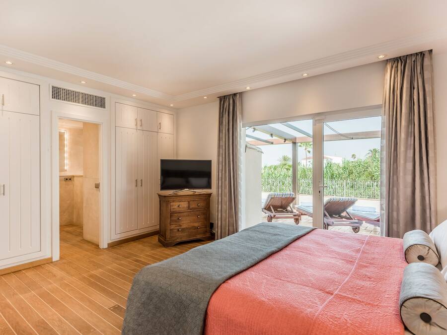 Un dormitorio con una cama grande, televisión y puerta corrediza de cristal que da a un área exterior con una silla y sombrilla.
