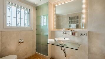 A bathroom with a glass shower enclosure, a glass sink on a glass shelf, a toilet, and a window with decorative glass panes.