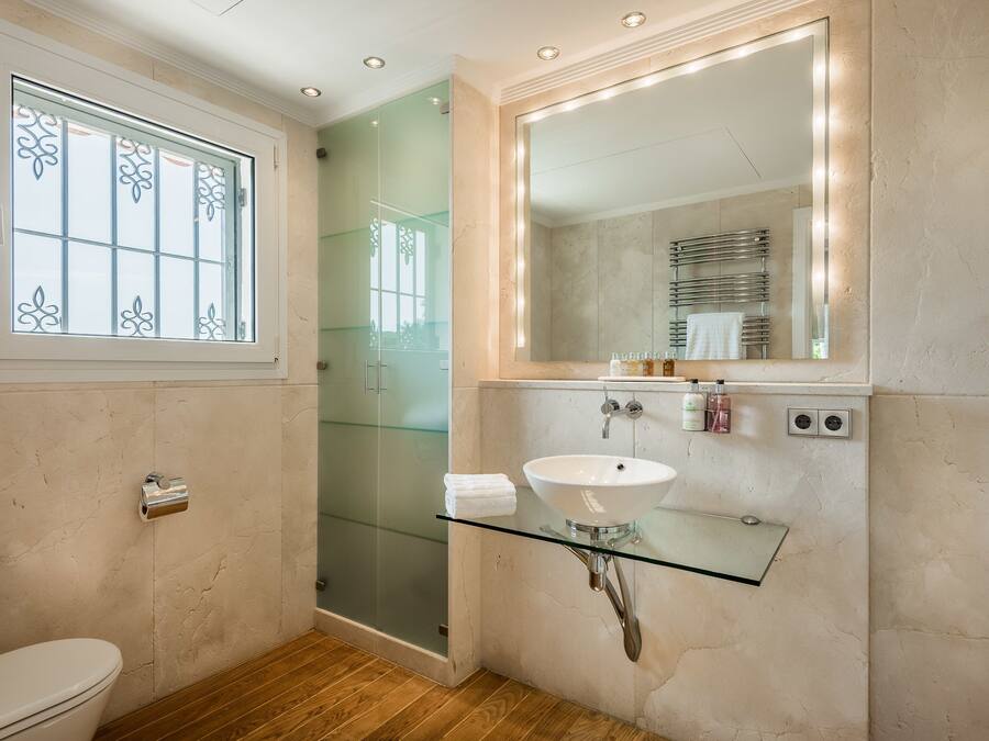 A bathroom with a glass shower enclosure, a glass sink on a glass shelf, a toilet, and a window with decorative glass panes.