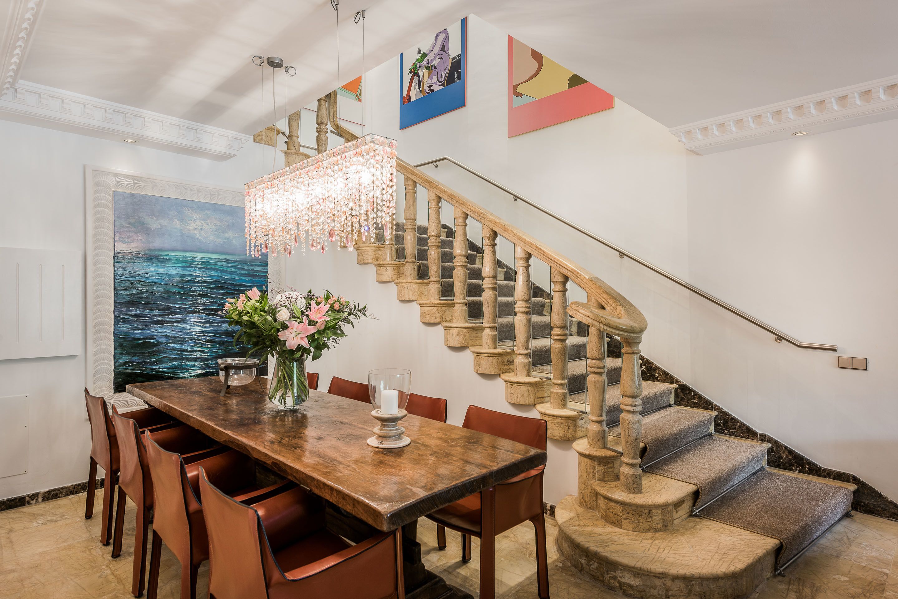 Un comedor con una mesa de madera, sillas y una araña. Una escalera con baranda de madera conduce a un piso superior.