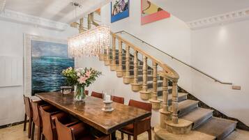 Un comedor con una mesa de madera, sillas y una araña. Una escalera con baranda de madera conduce a un piso superior.