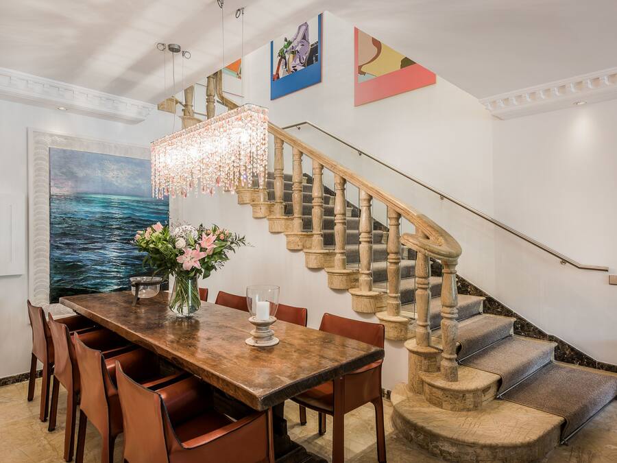Un comedor con una mesa de madera, sillas y una araña. Una escalera con baranda de madera conduce a un piso superior.