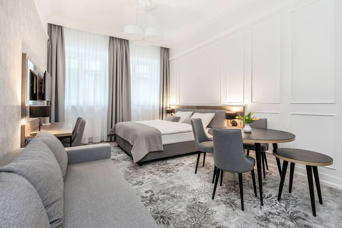 A modern hotel room with a bed, a sofa, a dining table, and a TV.