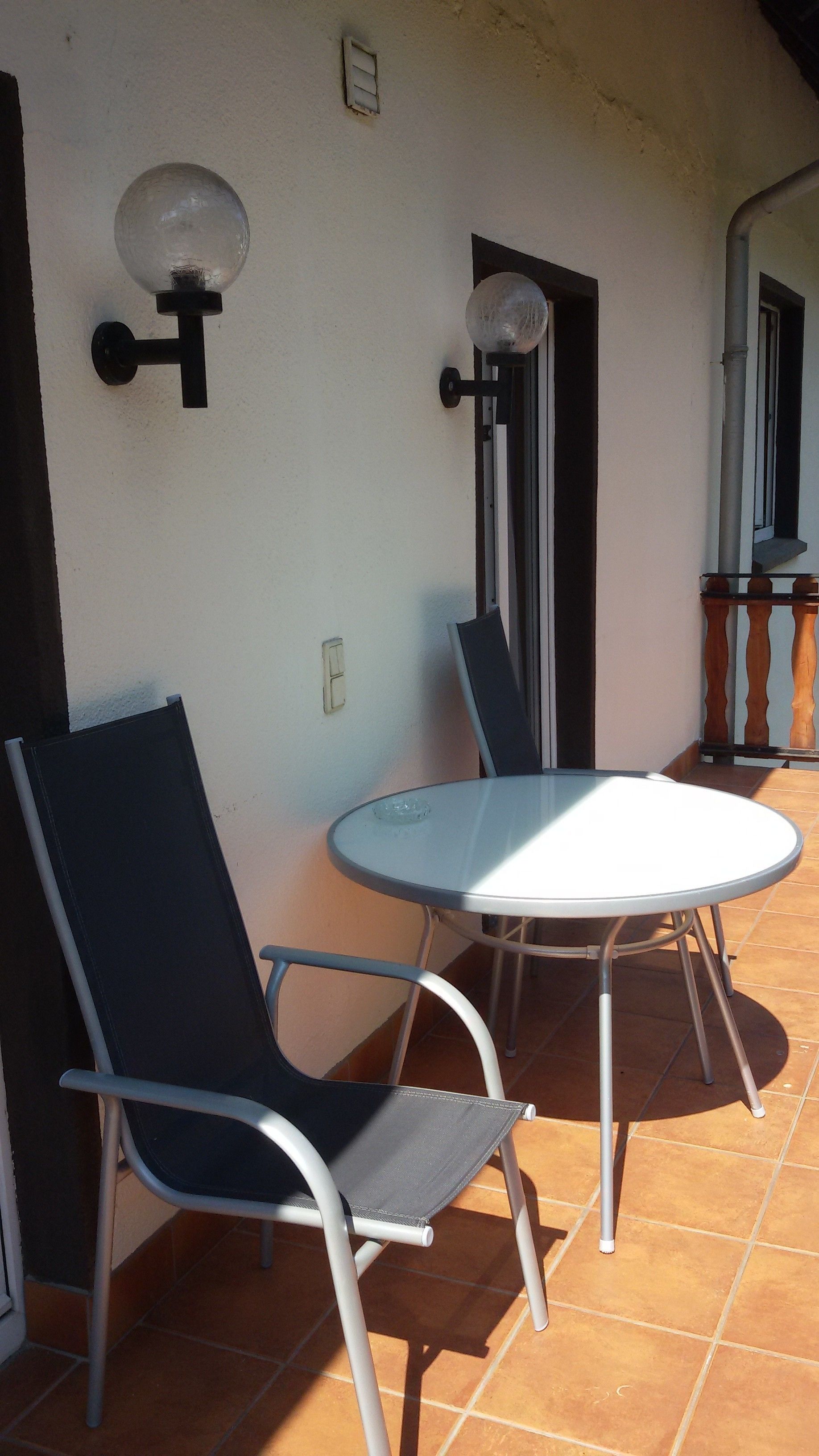 Une terrasse avec une table ronde et deux chaises, un luminaire fixé au mur et une fenêtre donnant sur l’extérieur.