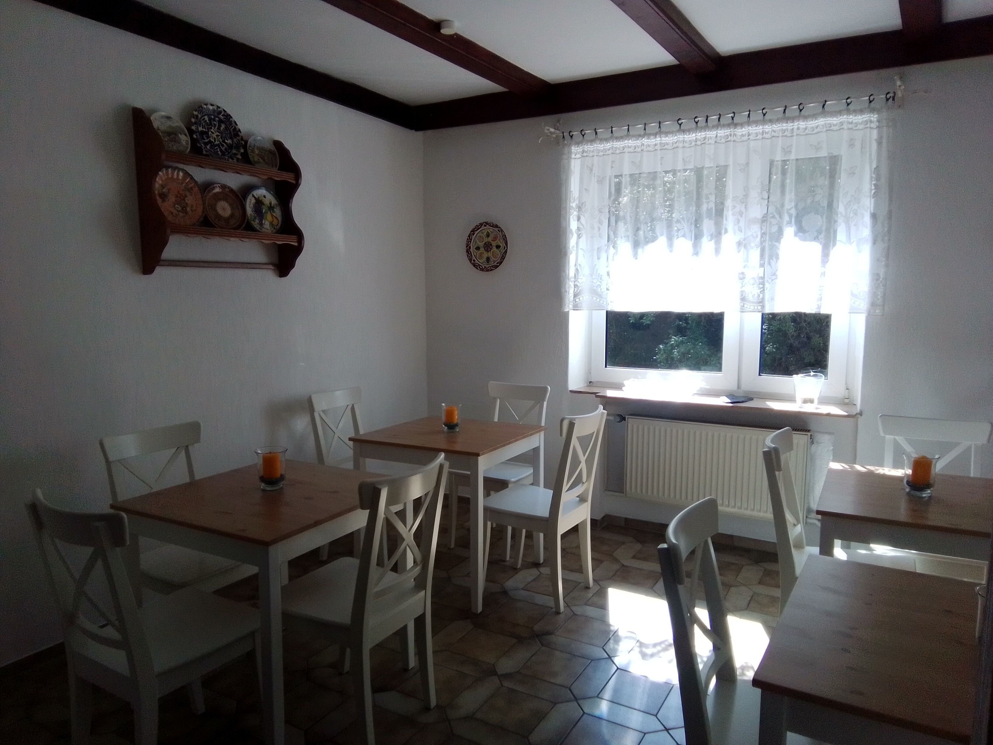 A dining area with wooden tables and white chairs, a window with a view of greenery, and a wall-mounted shelf with decorative plates.