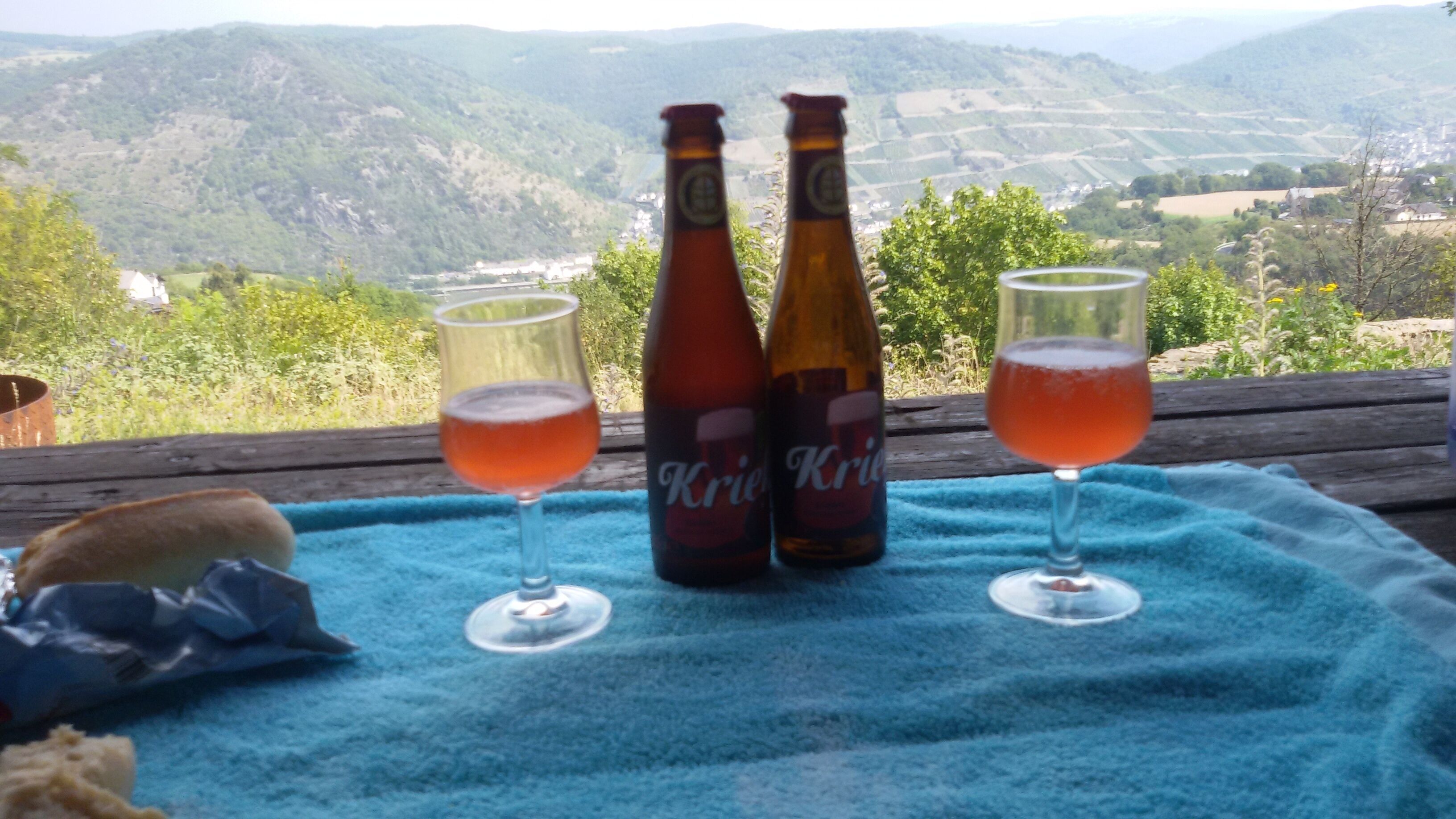 Zwei Kriez-Bierflaschen und zwei Biergläser auf einer blauen Decke, mit einer schönen Aussicht auf Hügel und Felder im Hintergrund.