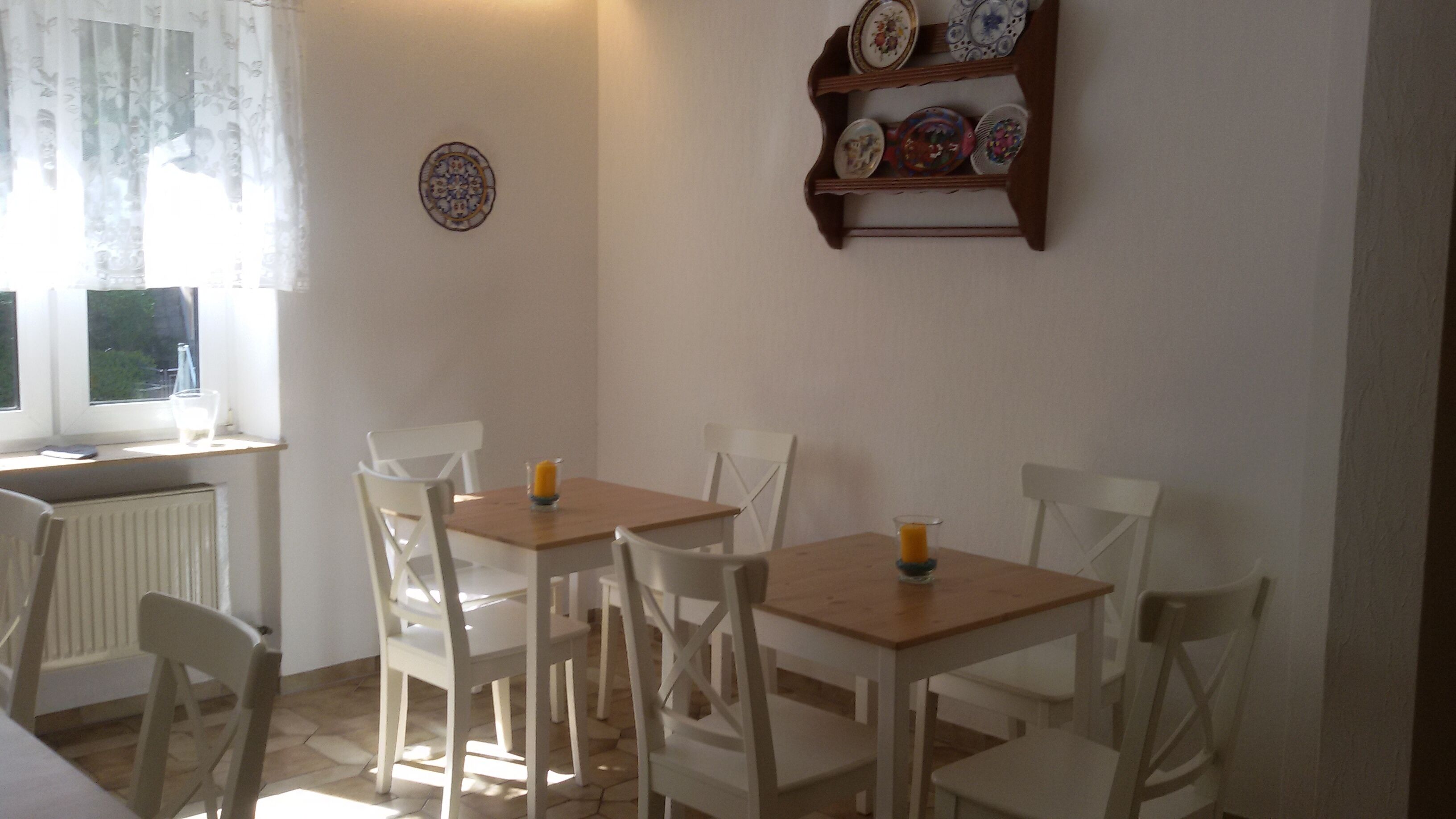 Een eetgedeelte met vier witte stoelen rond een houten tafel, een raam met dunne gordijnen, en een wandplank voor borden.