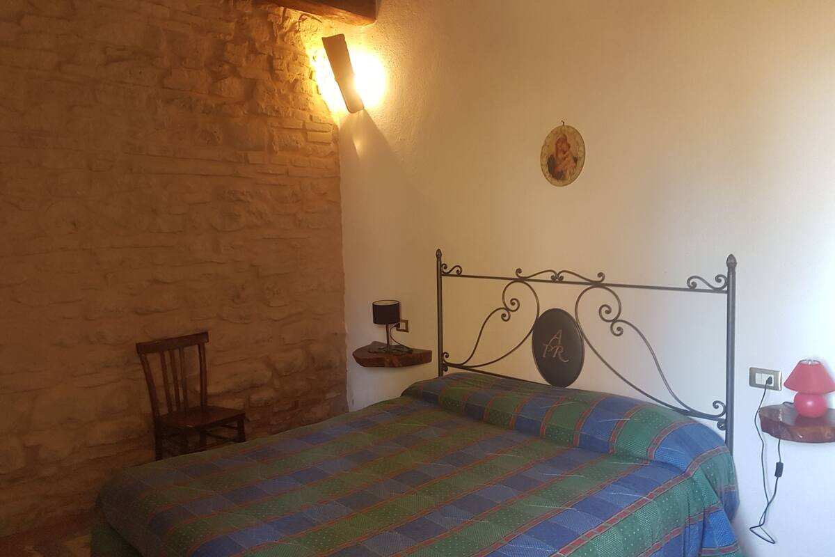 Ein Schlafzimmer mit Backsteinwand, einem Metallbettgestell, einer Steppdecke, einem Sessel, einer Wandlampe und einem kleinen Tisch mit Lampe.