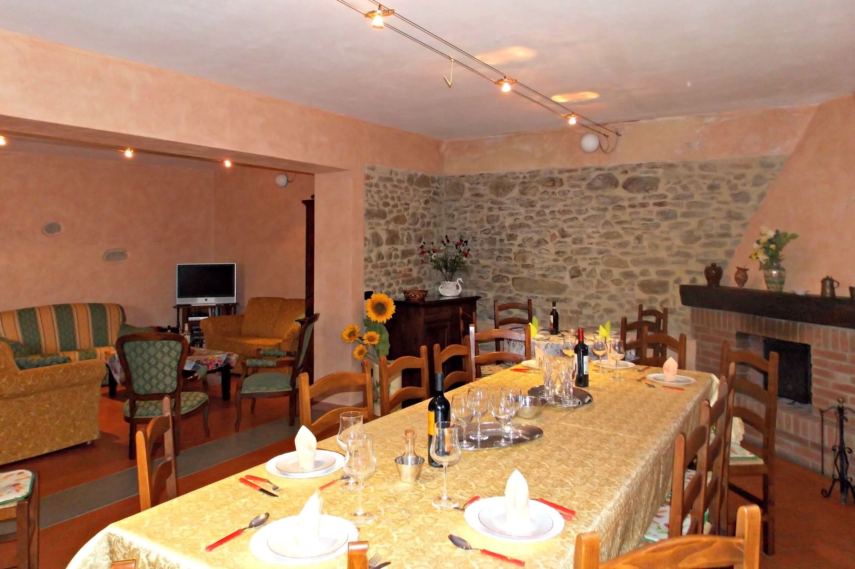 Un comedor con una larga mesa dispuesta para una comida, rodeada de sillas de madera. Se observa una pared de piedra y una chimenea al fondo.