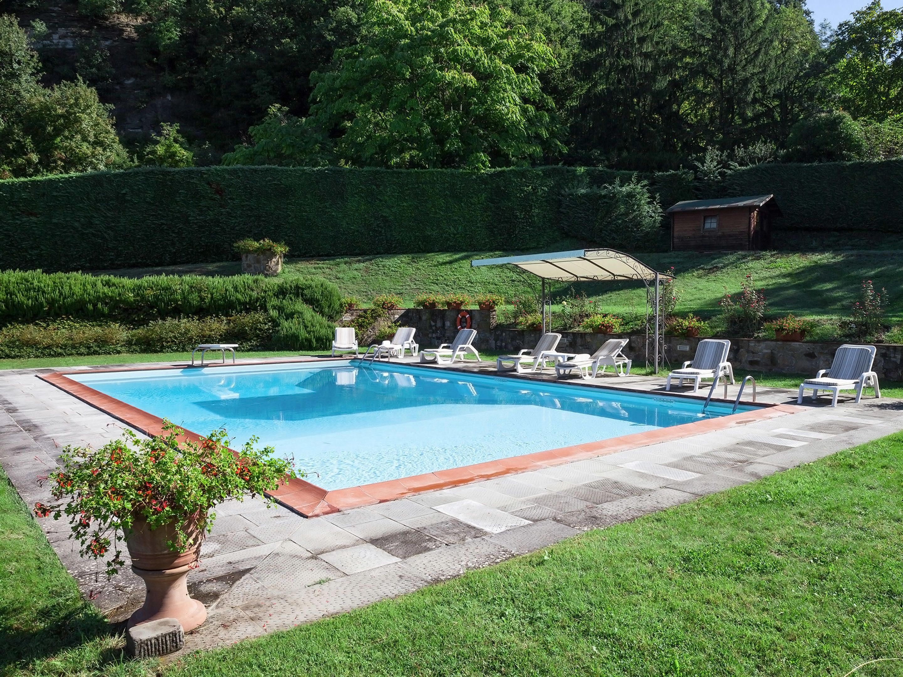 A rectangular swimming pool with a tiled edge, surrounded by a grassy area and a stone wall. There are several lounge chairs and a covered patio area with a metal frame and a white canopy.