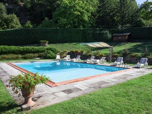 A rectangular swimming pool with a tiled edge, surrounded by a grassy area and a stone wall. There are several lounge chairs and a covered patio area with a metal frame and a white canopy. - Villa Eleonora (Dicomano)