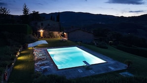 A rectangular swimming pool with a clear blue water surface, surrounded by a paved deck. There are lounge chairs aligned along the pool's edge, and a small table is placed near the pool's corner. The pool is illuminated by soft lighting, and the surrounding area includes a grassy lawn and a stone building in the background. - Villa Eleonora (Dicomano)