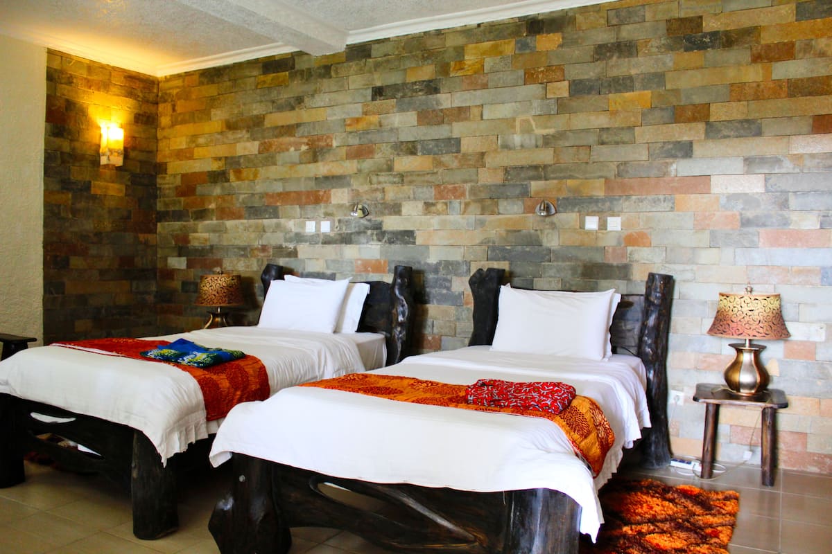 A hotel room with two beds, a stone wall, and a small table with a lamp.