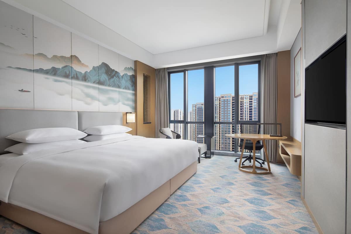 A modern hotel room with a large bed, a desk, and a view of the cityscape through large windows.