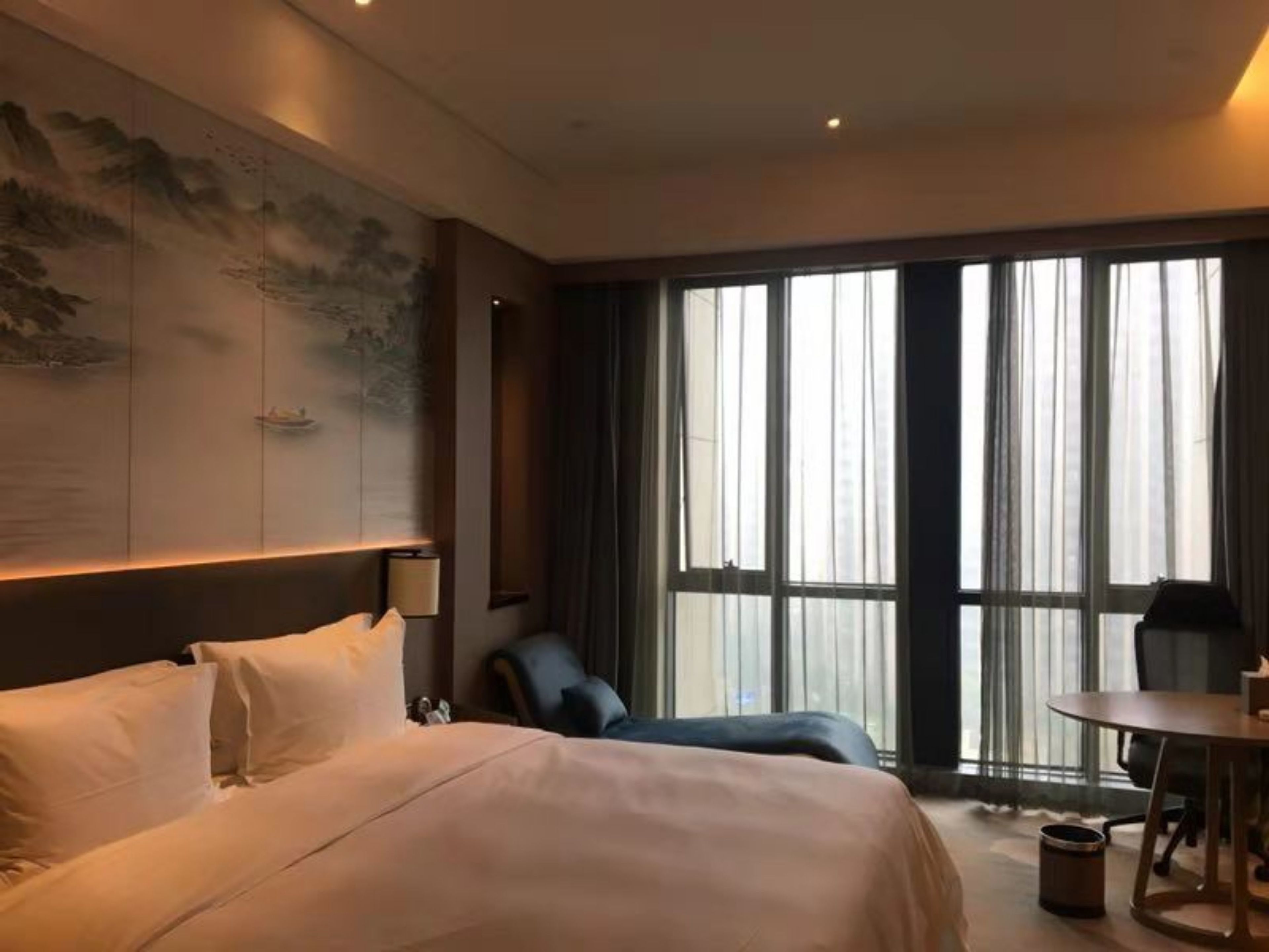 Habitación de hotel con cama, escritorio, silla, ventana grande con cortinas y una obra de arte en la pared.