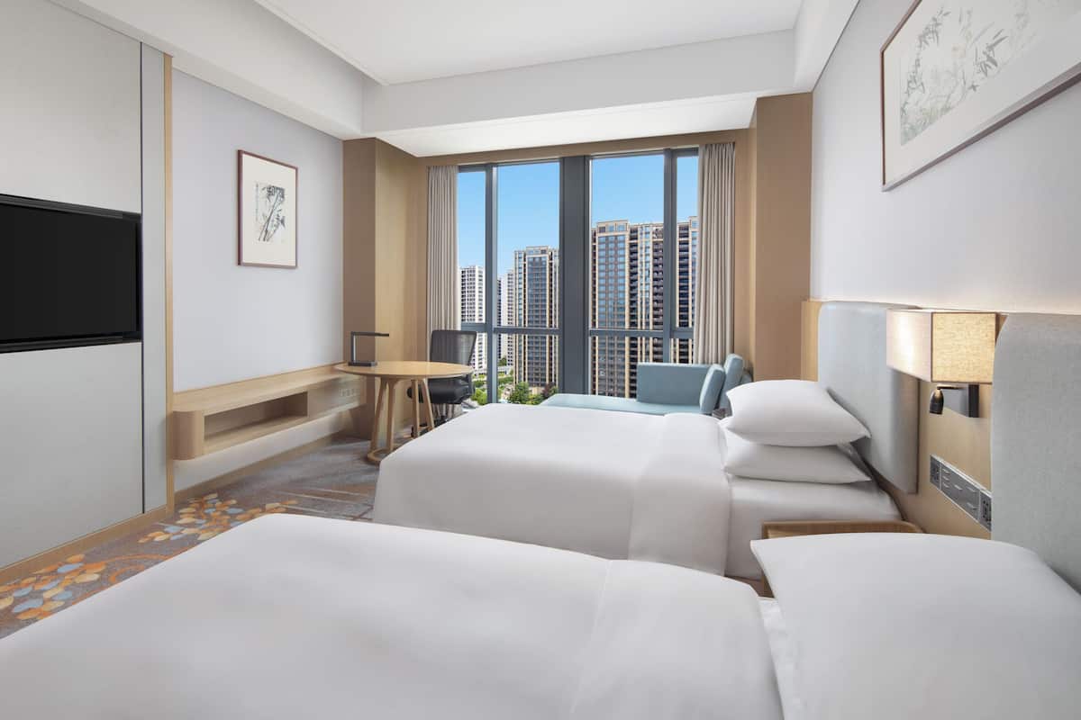 A hotel room with two beds, a desk, a chair, a TV, and a large window with a city view.
