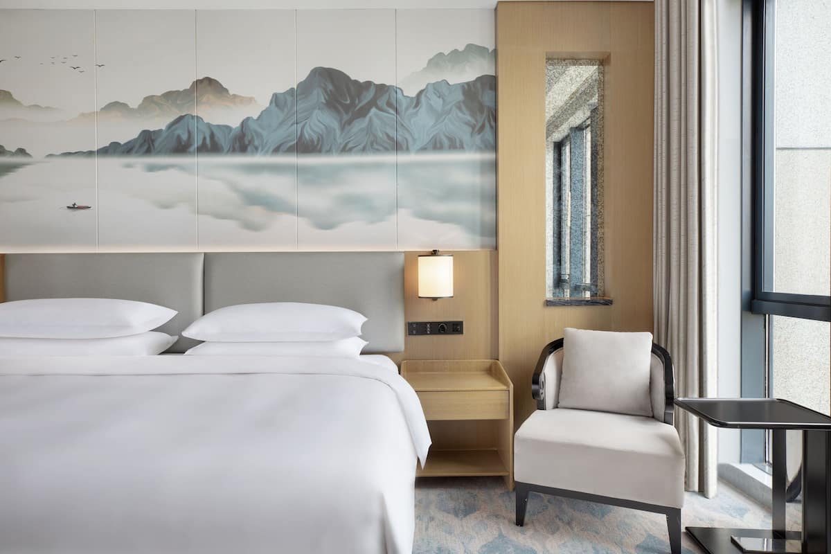 A hotel room with a large bed, a chair, a small table, a nightstand, and a wall-mounted lamp.