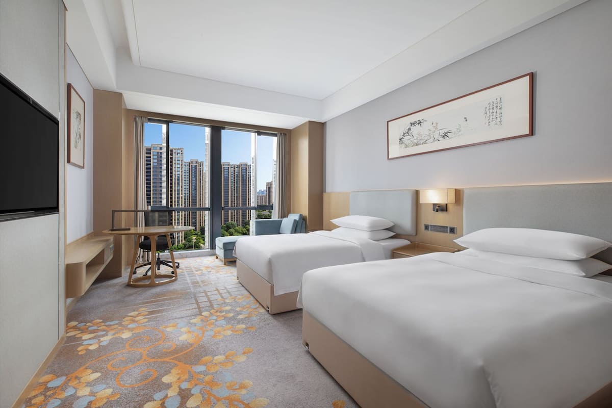 A hotel room with two beds, a desk, a chair, and a large window offering a city view.