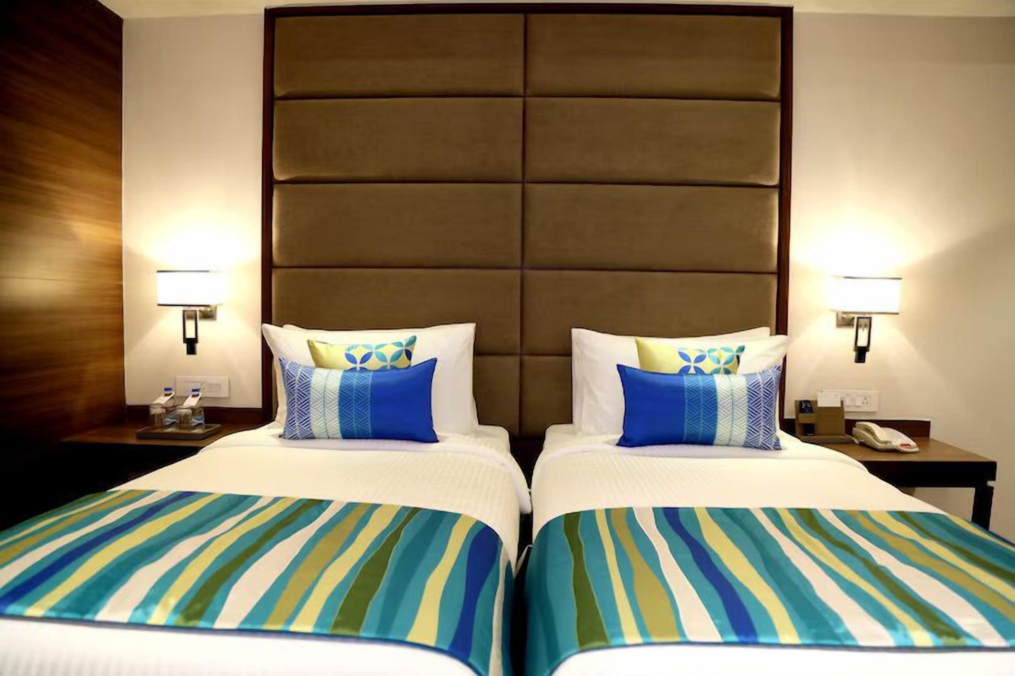 Een hotelkamer met twee bedden, elk voorzien van gestreept beddengoed en decoratieve kussens. Er staan nachtlampjes en een nachtkastje met een telefoon en een klein decoratief object.