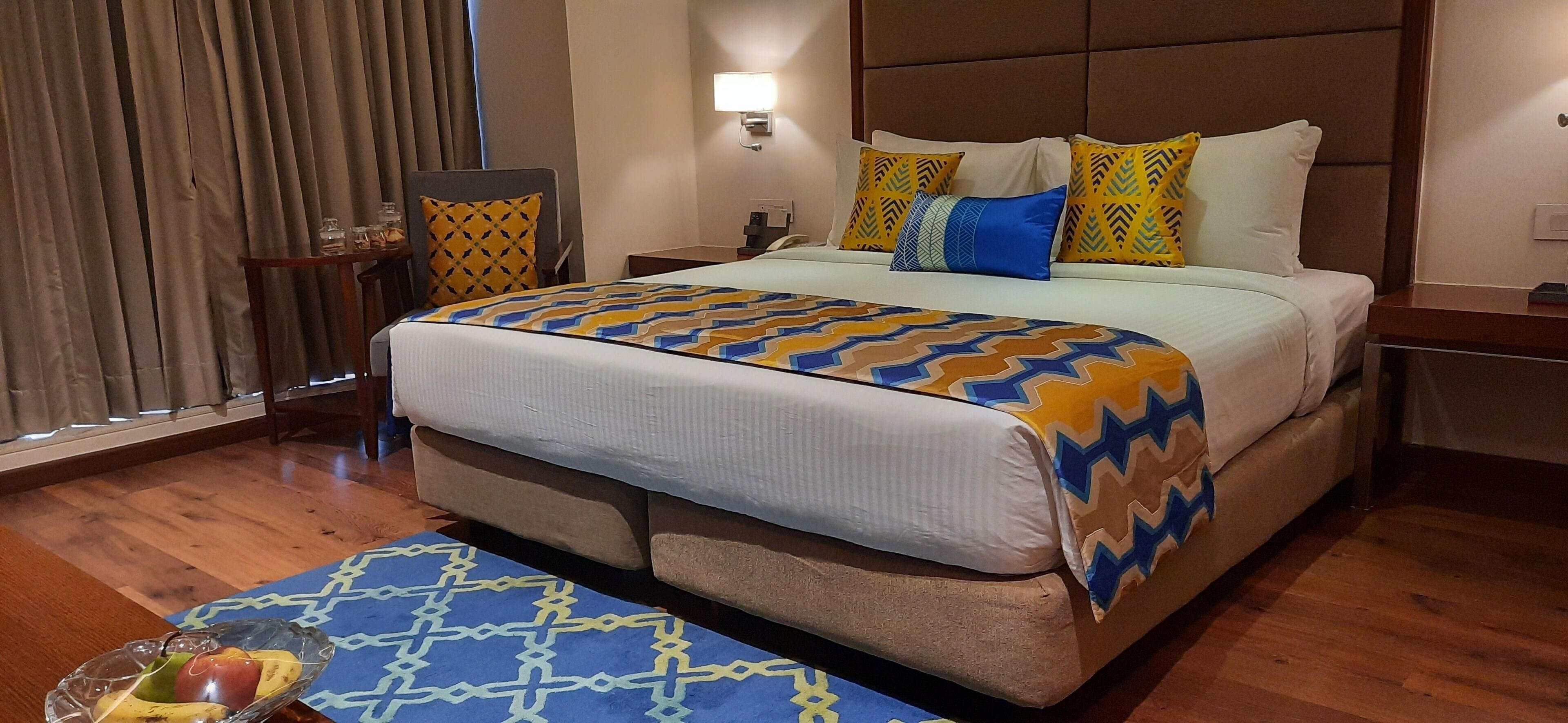 A hotel room with a large bed, a bedside table, a chair, a small table with a fruit bowl, and a window with curtains.