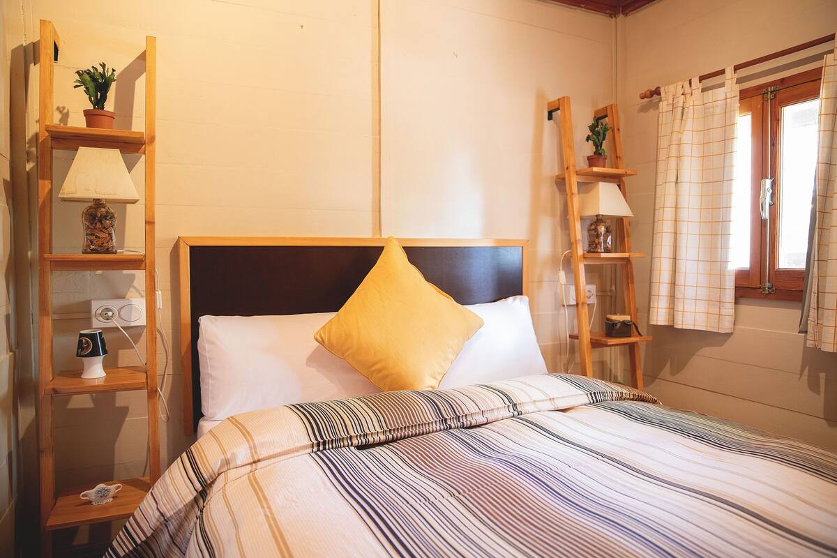 A neatly made bed with striped bedding, a wooden headboard, and a yellow pillow. There are wooden shelves with lamps and a potted plant on the left.