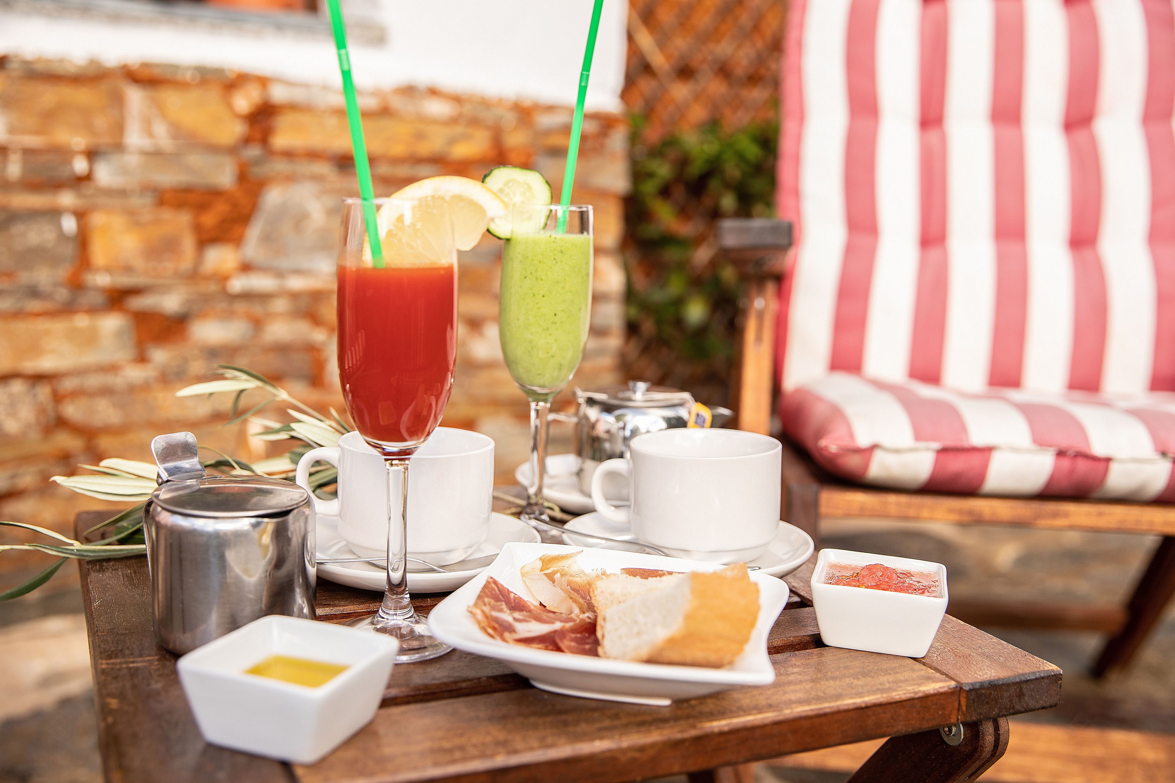 Une table en bois, dressée pour le repas, avec une carafe, des verres, un verre de boisson rouge, un verre de smoothie vert, du pain et un petit bol de sauce.