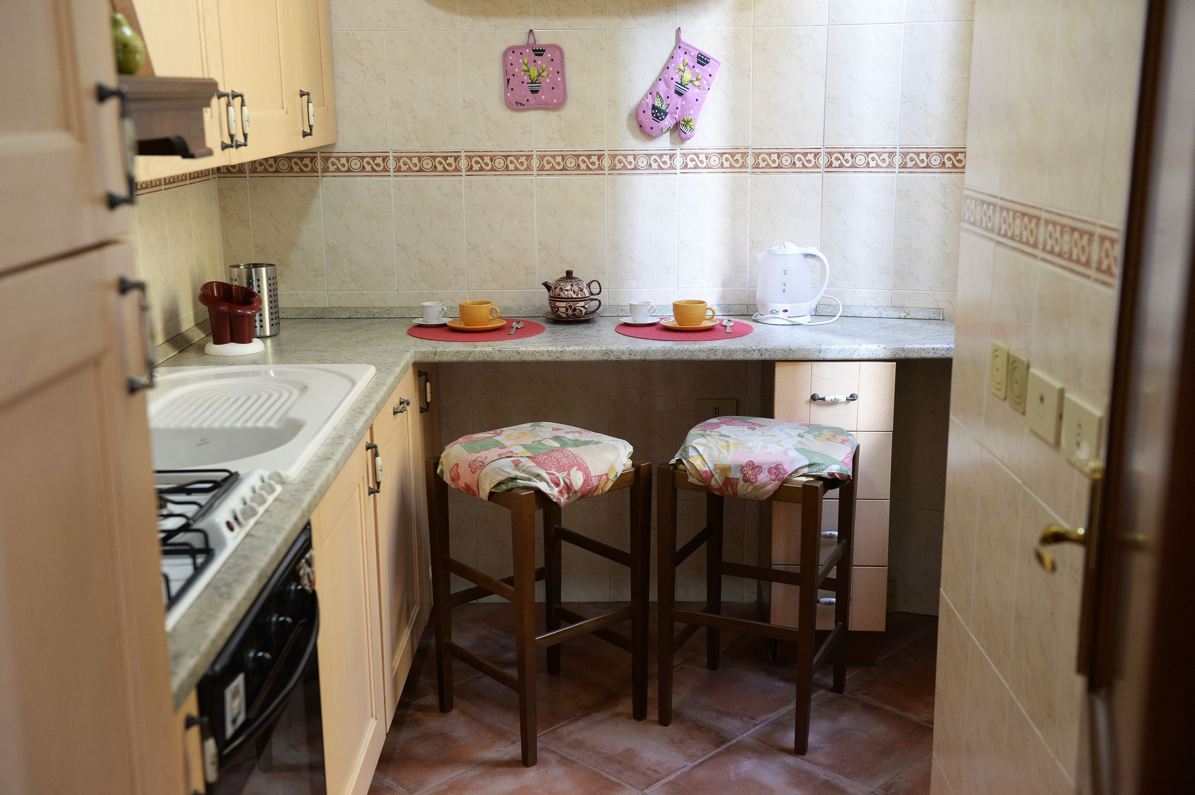 Una cocina con estufa a gas, dos taburetes tipo bar, cafetera y hervidor en la encimera.