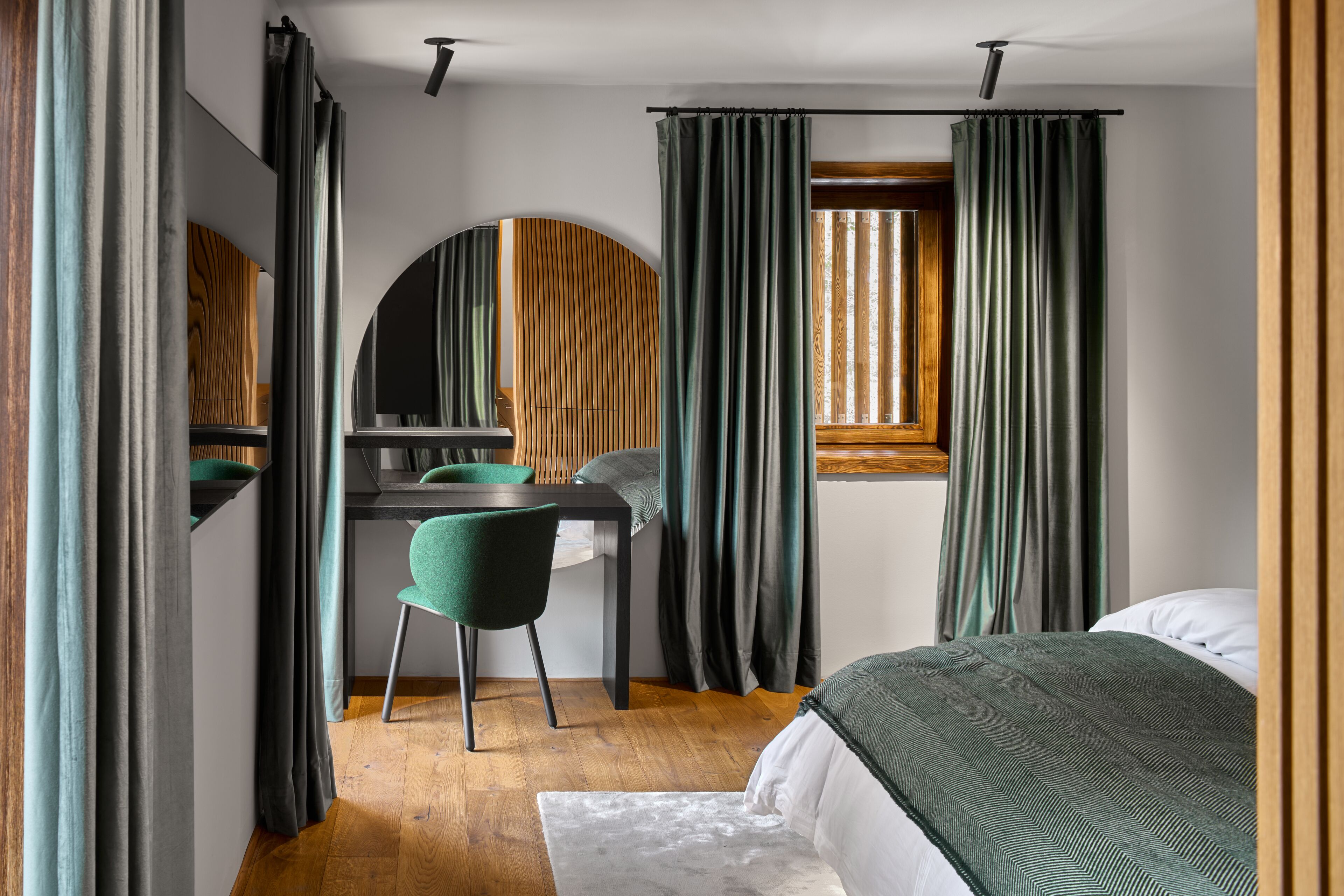 A bedroom with a bed, a desk, and a chair. There is a window with curtains, a mirror, and a wooden floor.