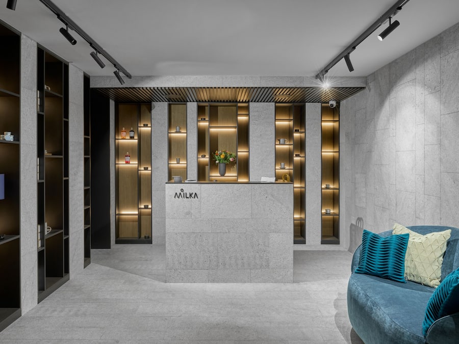 A modern reception area with a central desk labeled "MILKA," flanked by shelving units and a seating area with teal cushions.