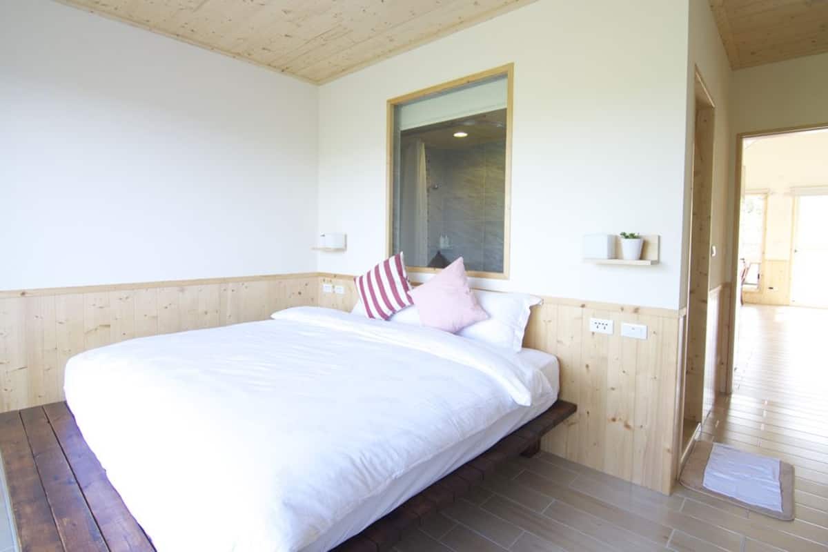 Een slaapkamer met een houten bedframe, wit beddengoed, een raam en een klein plankje met een plant.