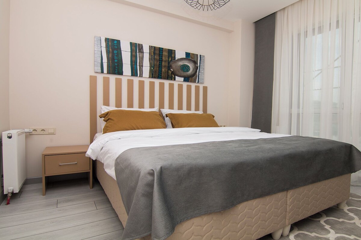 Ein modernes Hotelzimmer mit einem großen Bett, einem Nachttisch, einem Heizkörper und einem Fenster mit durchsichtigen Vorhängen.