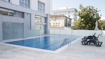 Una piscina al aire libre con agua azul cristalina, rodeada por un edificio moderno con grandes ventanales y suelo de baldosas.