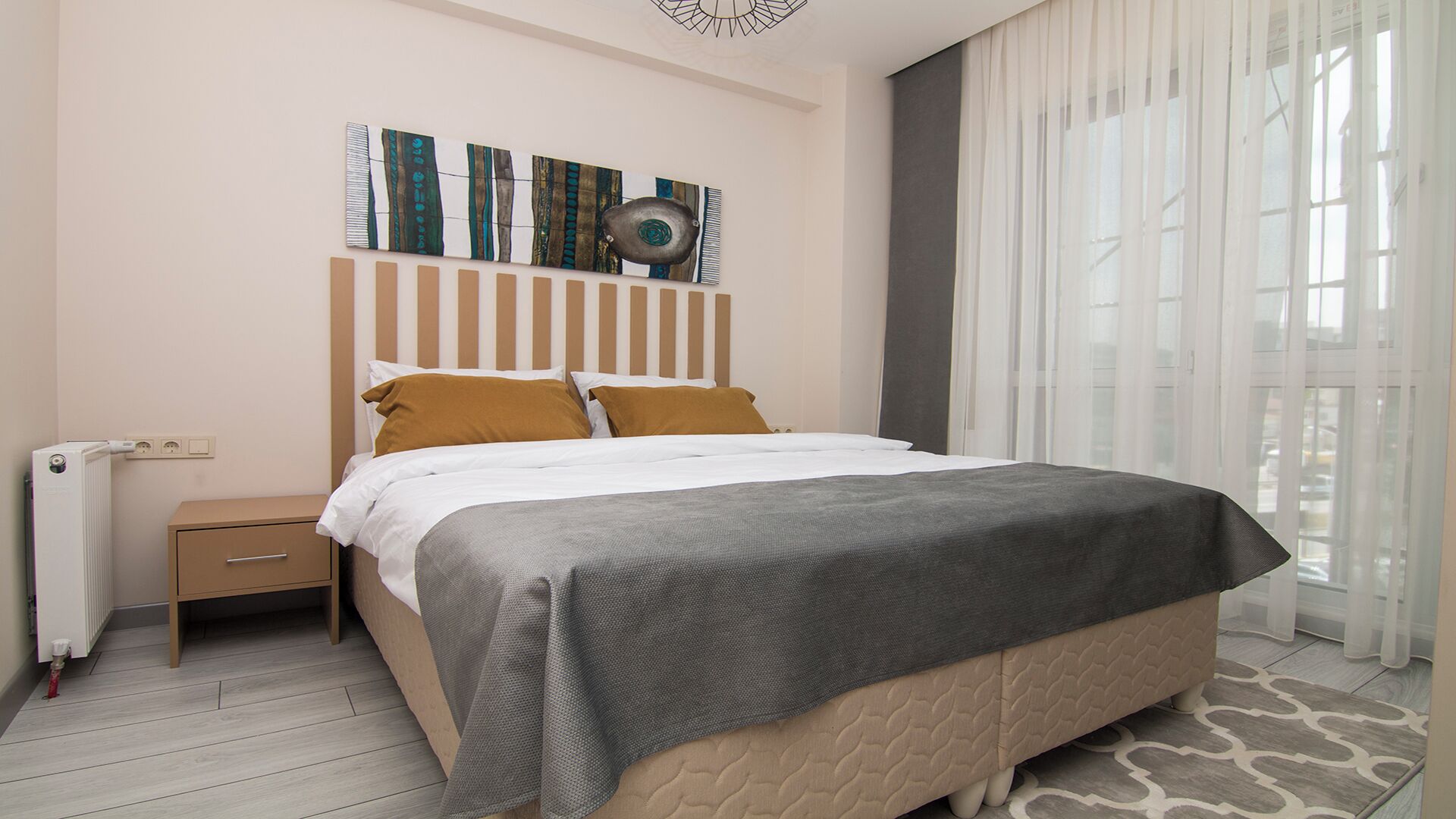 Une chambre d’hôtel moderne, équipée d’un lit, d’une table de chevet, d’un radiateur et d’une grande fenêtre avec des rideaux légers.