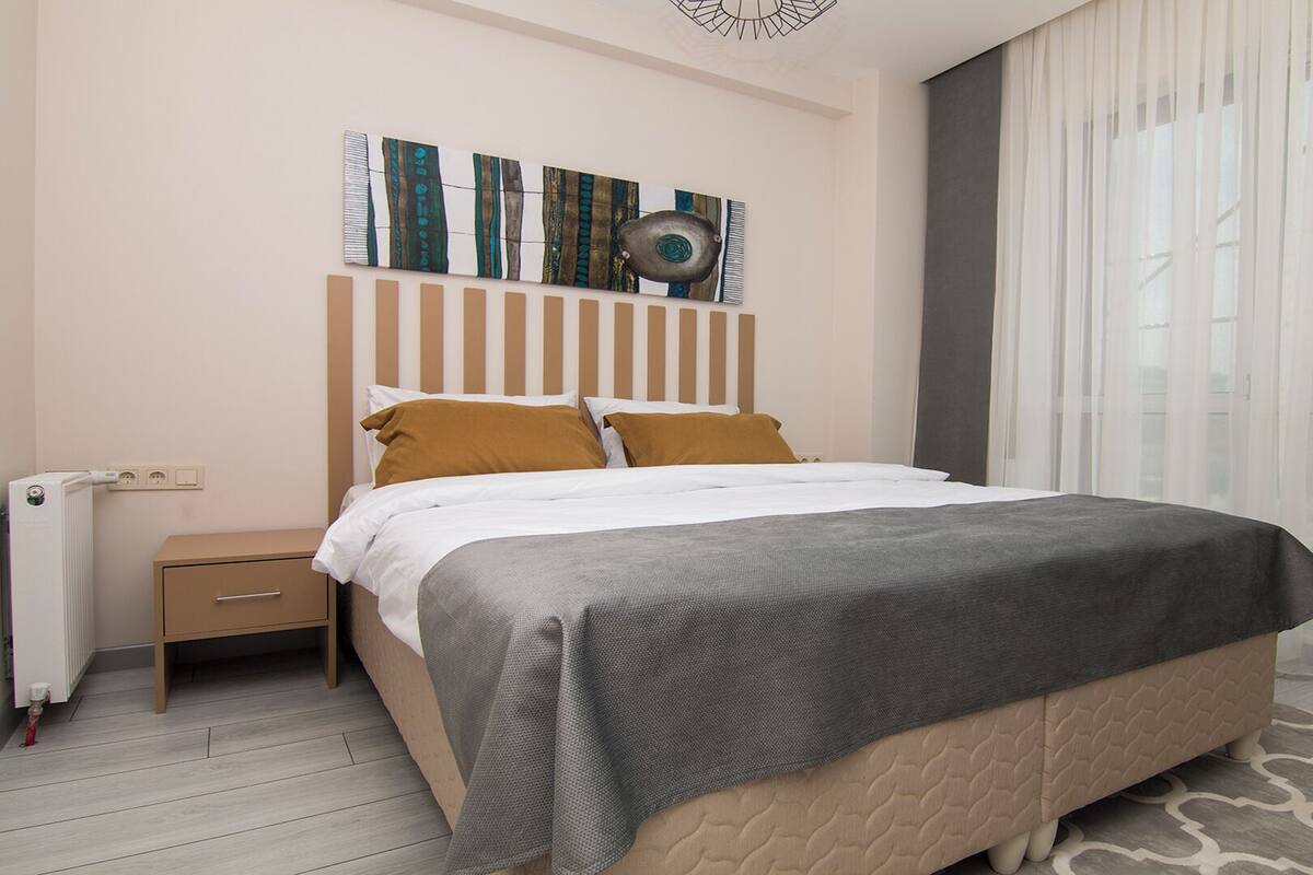A modern hotel room with a bed, a nightstand, a radiator, and a large window with sheer curtains.