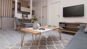 Un salon moderne comprenant un canapé, une table à manger avec des chaises, un téléviseur fixé au mur et un coin cuisine visible à travers une porte ouverte.