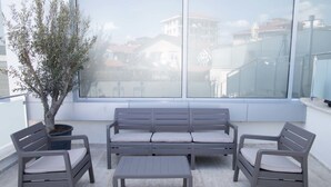 Un balcon avec un canapé gris, deux fauteuils et une petite table. On y trouve un arbre en pot et, au loin, une vue sur des immeubles.