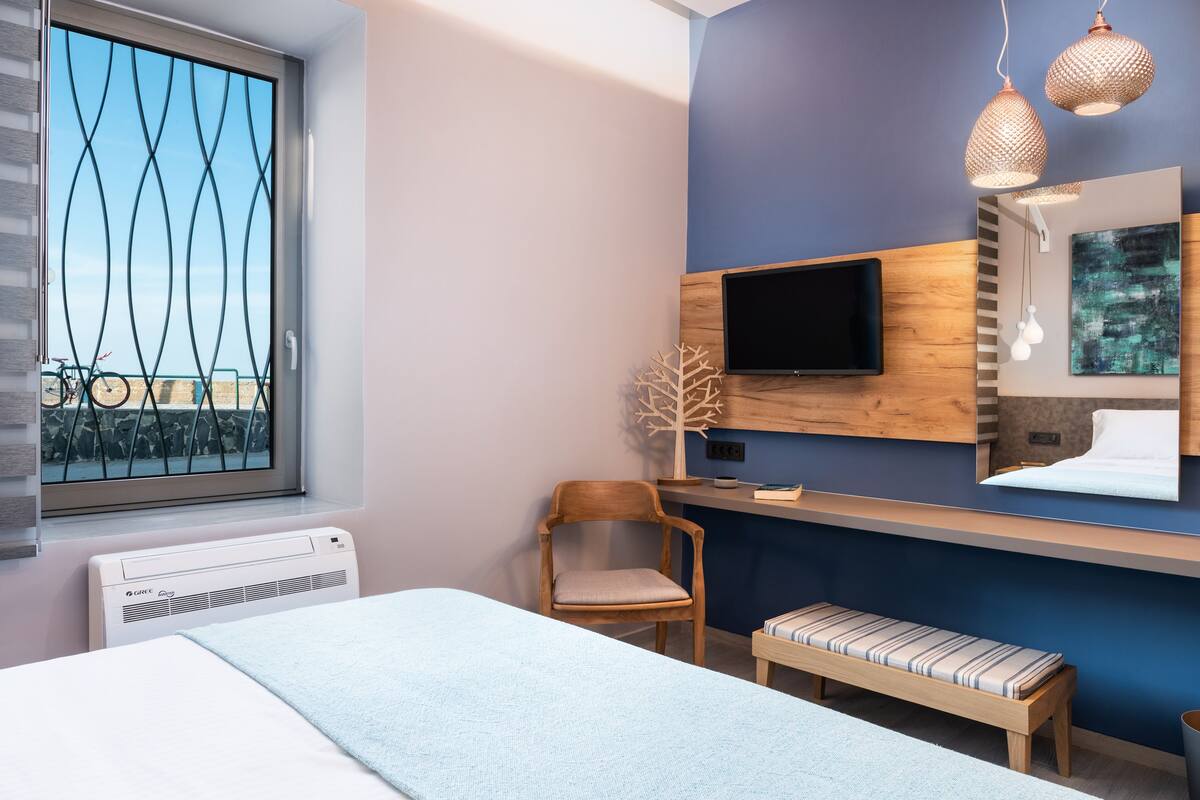 A modern hotel room with a bed, a wooden chair, a bench, a TV mounted on the wall, and a window with a view of bicycles.