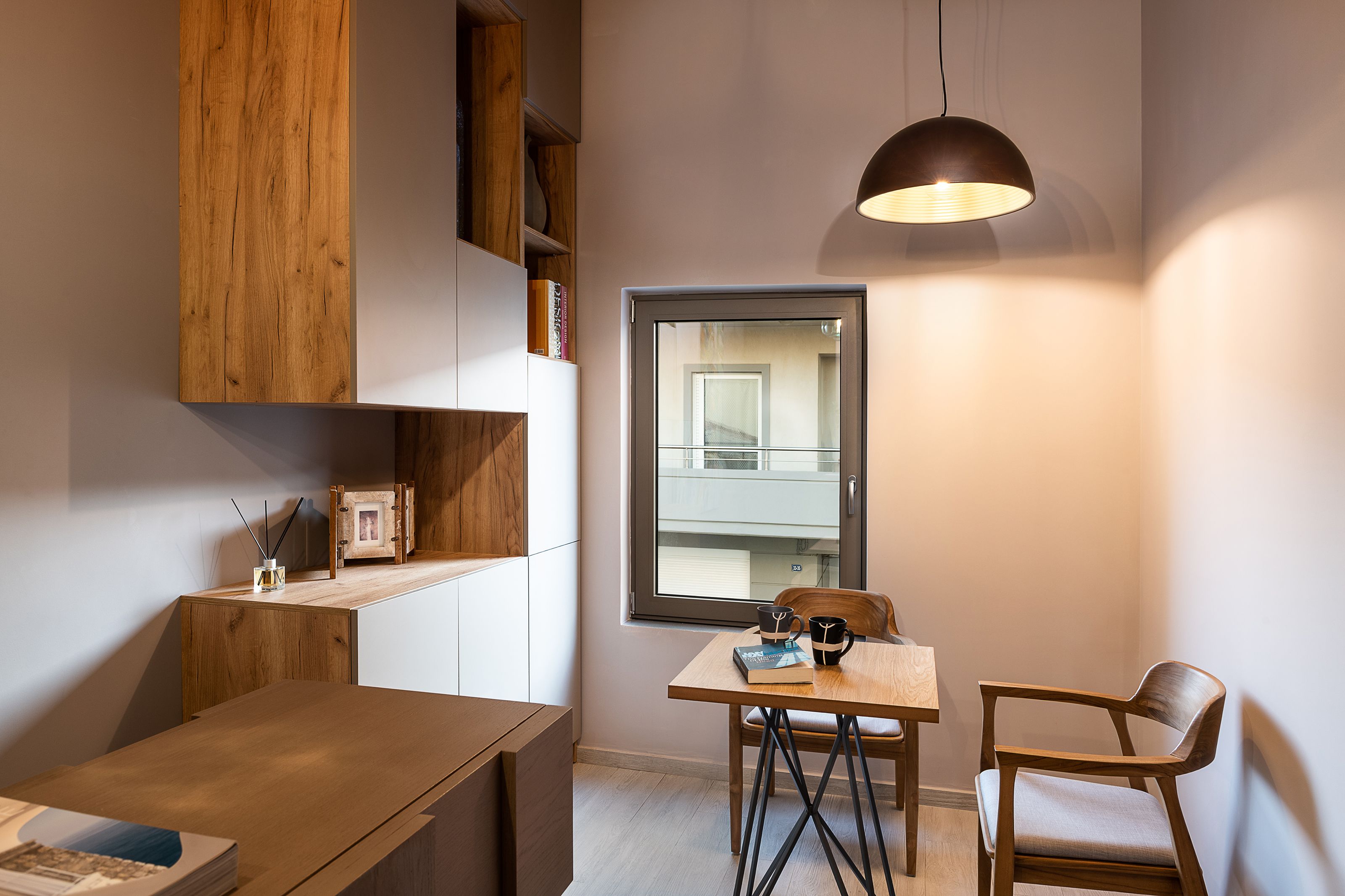 A compact room with a wooden table, a chair, a bookshelf, and a window with a view of a building.