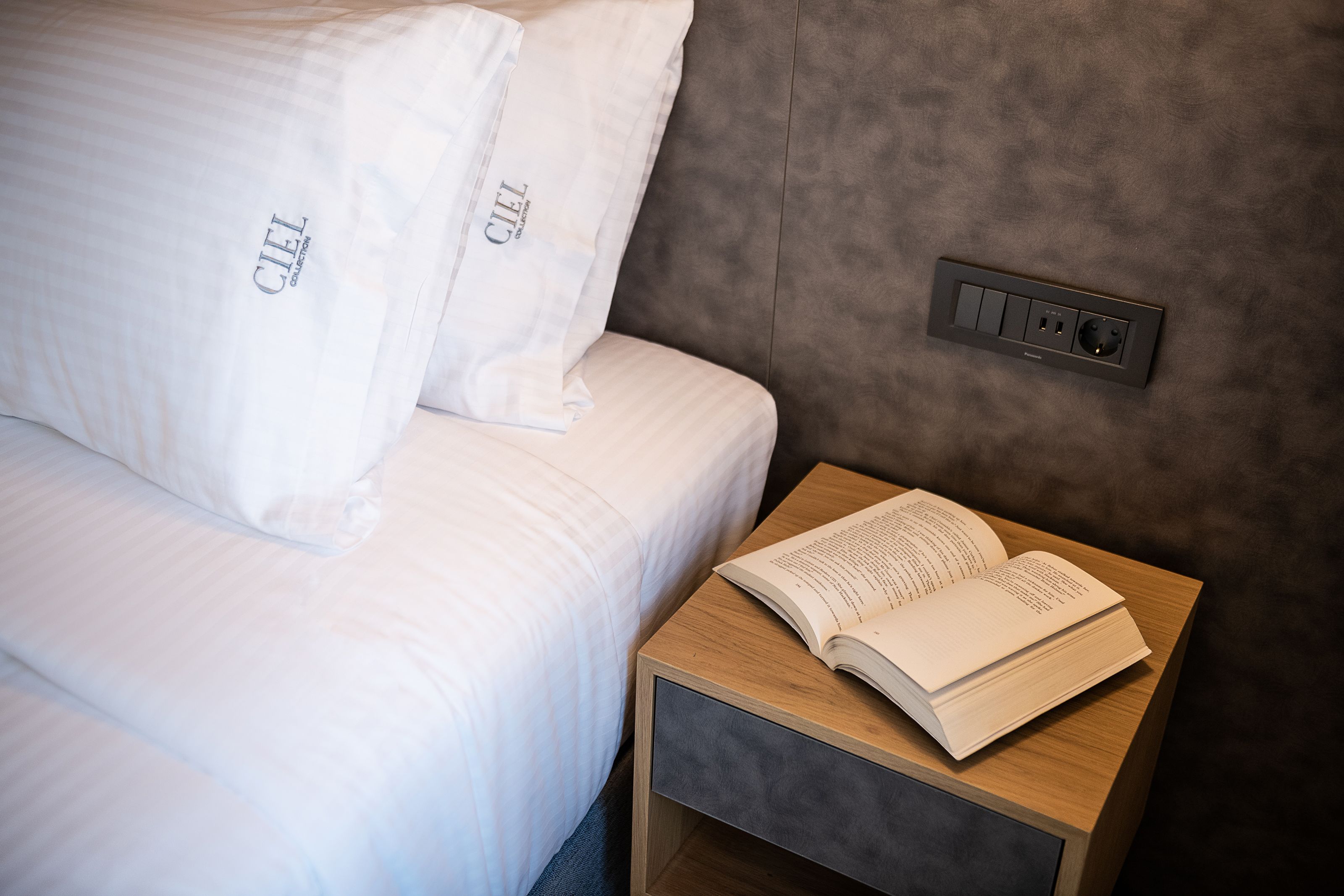 Una cama bien hecha con sábanas y almohadas blancas, una mesita de noche con un libro abierto y un enchufe en la pared.